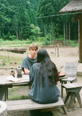 Tom and Nanao chatting in the outdoor dining area, Tom wears a cotton flax poplin folk top and wrapped pants, Nanao is wearing a folk twisted dress